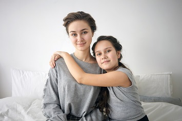 Perfect family. Portrait of cute 12 year old girl with long dark hair sitting on bed and hugging her beautiful young mother, smiling and looking at camera, spending weekend morning at home together